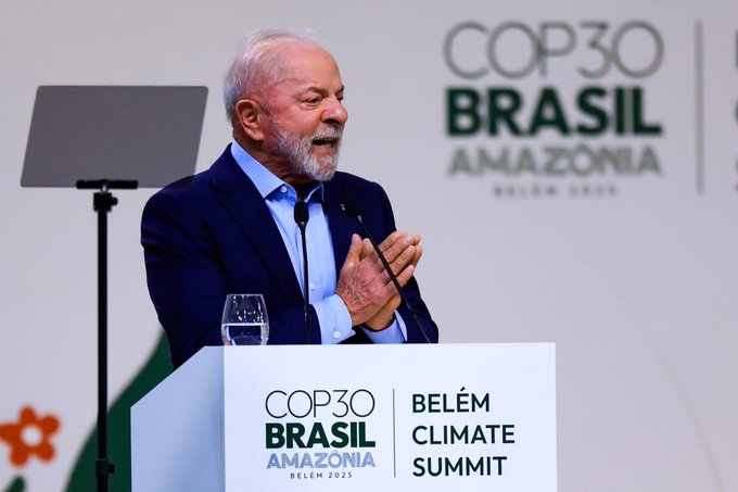 An elderly man with white hair and beard wearing a dark suit and light blue shirt stands at a podium speaking into a microphone, gesturing with clasped hands, positioned in front of a large backdrop displaying the COP30 Brasilia Amazonia Belem Climate Summit 2025 logo in green and white colors, with additional signage for COP30 Belem on the podium and a glass of water nearby, set in a conference hall with neutral background.