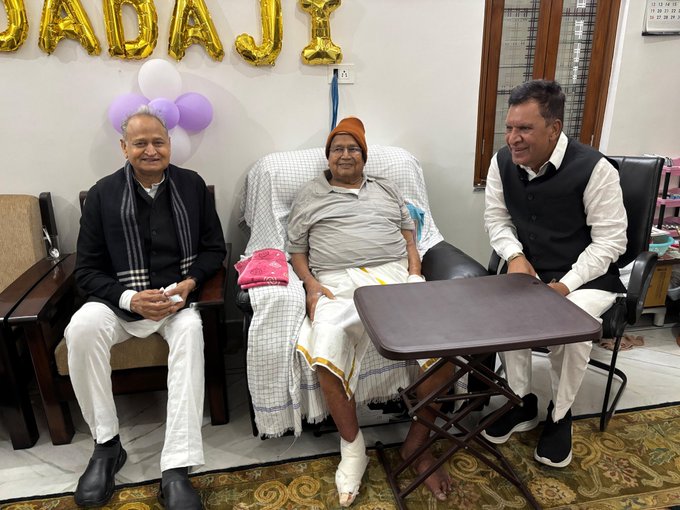 Three elderly men seated in a living room with beige walls and a window. Left man in black sweater and white pants holds hands together on lap. Center elderly man with white beard wears beige cap and light shirt, feet bare with right foot bandaged white, sitting on cushioned chair with white sheet. Right man in white vest and pants smiles. Small brown table between center and right chairs. Purple and gold balloons and Hindi text banner on wall behind. Pink pillow on left chair arm. Brown leather chair left, beige right. Floral patterned floor rug.