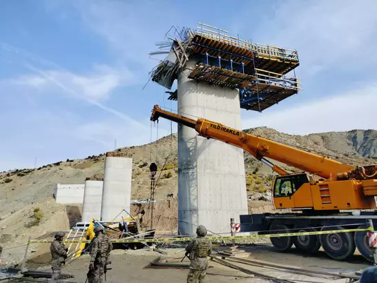 Construction site at a viaduct in a hilly area with multiple concrete pillars some under construction orange Liebherr crane lifting materials near a collapsed scaffolding section on the central pillar workers in camouflage uniforms standing nearby inspecting the area scattered debris and equipment on the ground blue sky with clouds in background