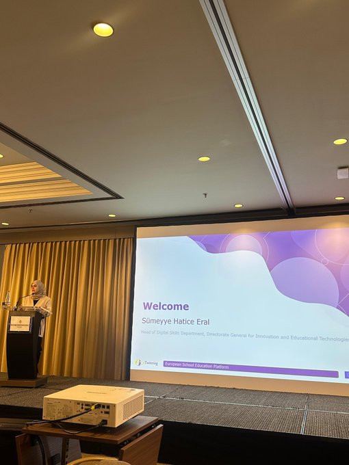 First image shows a male speaker in a suit standing at a podium in a modern conference room with yellow lighting and beige walls addressing an audience with a large screen displaying a welcome slide for eTwinning Central Support Service event on educational technologies. Second image depicts a female presenter in white blouse leading a roundtable discussion with diverse group of women including some wearing headscarves seated around a table with water bottles notebooks and a screen showing colorful graphics in a brightly lit room with large windows. Third image captures a panel discussion with four participants including women and men in professional attire seated on chairs facing a screen with purple-themed slide titled Leadership for Change what does it mean today featuring a lightbulb icon in an elegant room with gold curtains and chandeliers. Fourth image illustrates a female speaker in red top presenting near a projector screen displaying video footage of children with a diverse audience of adults some in headscarves seated at round tables with water bottles in a formal conference hall with beige curtains and wooden podium.