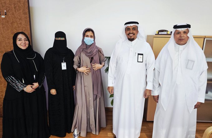 Group of five people standing in an office setting with wooden paneling and shelves in the background. Two women on the left wear black abayas with embroidered details and name tags, one smiling with headscarf, the other in full niqab and mask. Two men in the center and right wear white thobes and red-and-white checkered headscarves, one with a name tag, all appearing professional and engaged in a formal meeting context.