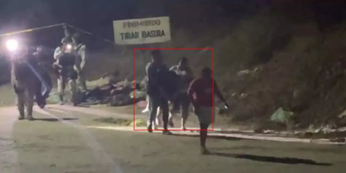 Nighttime scene on a rural road with a sign reading Tribasora illuminated by vehicle lights. Several armed men in dark clothing stand around three prone bodies on the ground near the sign. A red pickup truck is parked nearby with its lights on. The area is surrounded by bushes and rocks under a dark sky.