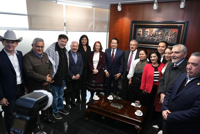 Group of ten adults mostly men in suits and women in dresses and blazers standing posed in a formal room with wooden walls and a large abstract painting of historical figures on the wall a low wooden table with white cups and saucers in front a tripod camera on the floor to the side one man wears a cowboy hat others have varied hairstyles and accessories including ties and jewelry the setting includes overhead lights and beige curtains