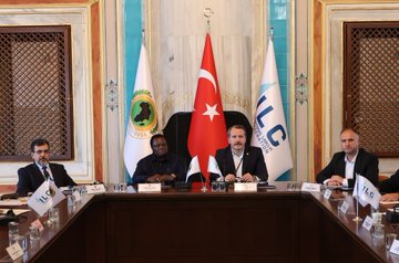 First image shows two men seated at a conference table with nameplates reading Francis Atwoli and Ali Yalcin, wearing suits and microphones, in front of flags including Turkish, Guinean, and ILC banners in an ornate room with blue and white decor. Second image depicts a large group of suited men and women around an oval wooden table in a grand hall with arched windows and blue tiled walls, discussing with documents and water glasses. Third image features four men in suits at a table with flags of Turkey, Guinea, and ILC, nameplates, and microphones in a room with intricate blue and gold patterns. Fourth image shows a diverse group of professionals seated around a long wooden table in a historic room with projector screen displaying text in Turkish and English about the meeting, chairs, and documents.