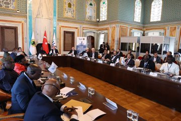First image shows two men seated at a conference table with nameplates reading Francis Atwoli and Ali Yalcin, wearing suits and microphones, in front of flags including Turkish, Guinean, and ILC banners in an ornate room with blue and white decor. Second image depicts a large group of suited men and women around an oval wooden table in a grand hall with arched windows and blue tiled walls, discussing with documents and water glasses. Third image features four men in suits at a table with flags of Turkey, Guinea, and ILC, nameplates, and microphones in a room with intricate blue and gold patterns. Fourth image shows a diverse group of professionals seated around a long wooden table in a historic room with projector screen displaying text in Turkish and English about the meeting, chairs, and documents.