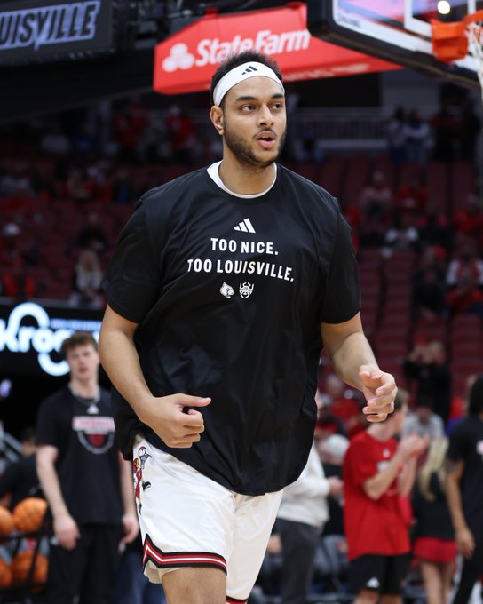 A male basketball player with a headband stands on an indoor court during a game, wearing a black Adidas t-shirt printed with Too Nice To Louisville text and logo, white shorts with red and black stripes, and red and white socks, surrounded by blurred spectators in the stands and a visible scoreboard with State Farm branding above.