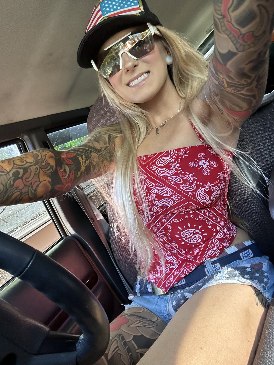 Woman with long blonde hair and extensive arm tattoos takes a selfie in a car interior, wearing mirrored sunglasses, American flag trucker hat, red bandana bandeau top, and cutoff jean shorts. She smiles while holding her phone arm extended. Steering wheel and seat visible in background.