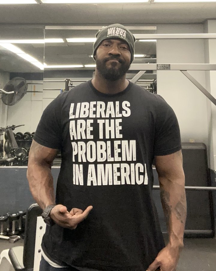 A muscular Black man with a beard and goatee wearing a black beanie stands in a gym with exercise equipment in the background. He wears a black t-shirt with white text reading LIBERALS ARE THE PROBLEM IN AMERICA. He flexes both arms outward while wearing gray gym shorts and a watch on his left wrist. The setting includes a fan, mirrors, and weights.