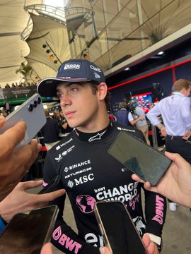 A young man with short dark hair wearing a navy blue cap with white BRB logo and a black racing suit featuring sponsor patches including Sparco collar, Binance, MSC, and BWT logos, surrounded by multiple hands holding black smartphones towards his face in a crowded press area under a white tent structure with green plants and blurred crowd in the background.