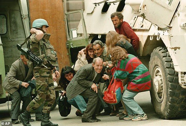 A historical color photograph depicting UN peacekeepers in camouflage uniforms and blue helmets, armed with rifles, escorting a group of civilians including women and children dressed in 1990s civilian clothing (e.g., colorful sweaters, handbags) near green military trucks and white armored personnel carriers in an urban street setting scarred by conflict, with civilians appearing anxious and huddled.