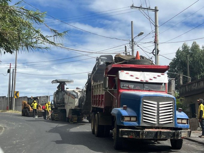 Road construction scene with a blue dump truck loaded with asphalt positioned behind a white paving machine spreading material on the street surface workers in yellow vests and helmets stand nearby directing operations orange traffic cones mark the work area power lines and trees line the roadside under a partly cloudy sky