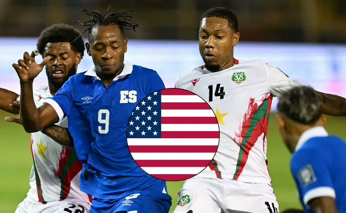 Several male soccer players in athletic uniforms compete on a green field during a match. One player in a blue jersey with number 9 and El Salvador flag emblem reaches for the ball while another in a white jersey with number 14 and Surinam flag emblem defends nearby. Additional players in similar uniforms are positioned around them engaging in the play. American flags are overlaid on the image.