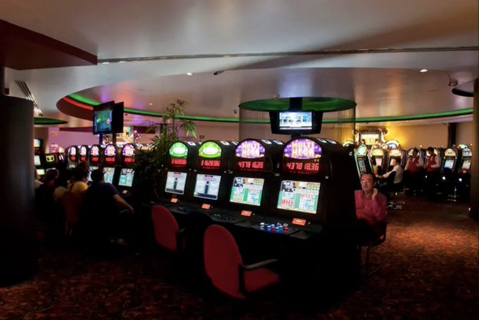 Interior of a Mexican casino with multiple rows of illuminated slot machines displaying various games and screens, green and yellow lighting accents on walls and ceiling, several patrons seated at machines playing, potted plants and large TV screens mounted above, red chairs and carpeted floor visible.