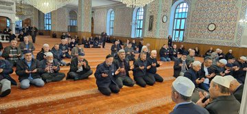 First image shows group of men in traditional clothing seated on prayer rug in ornate mosque interior with chandeliers, mihrab niche, and geometric wall patterns. Second image depicts larger gathering of men kneeling in prayer facing mihrab in similar decorated hall with windows and rugs. Third image captures men in white caps and suits seated and gesturing during event near mihrab and wooden panels. Fourth image illustrates men praying with hands raised towards speaker on platform beside columns and clock in mosque setting.