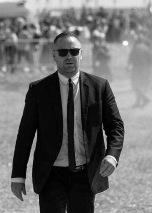 Black and white photograph of a middle-aged man with short hair, wearing sunglasses, a dark suit, white shirt, and black tie, walking outdoors toward the camera with a serious expression; blurred background shows a crowd of people behind metal barriers, security personnel, and event lighting, suggesting a public rally or march.