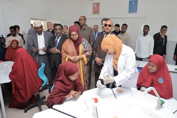 First image shows a group of men and women in formal attire including suits, traditional robes, and headscarves standing together cutting a red ribbon in front of a building entrance with blue walls, balloons, and a Maarif sign. Second image depicts women and girls in headscarves and modest clothing gathered around a table in a classroom with Turkish and Somali flags on the wall, one woman in a lab coat pouring liquid from a beaker into a test tube during a science experiment. Third image features a diverse group of men and women in suits, military uniforms, and traditional attire standing and sitting around desks with books and papers in a room with Turkish and Somali flags and a whiteboard. Fourth image displays several men and women in formal and traditional clothing standing around a table covered with stacks of colorful childrens books in a room with blue doors and windows.