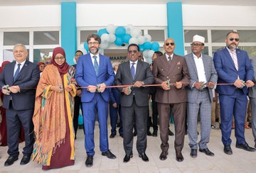 First image shows a group of men and women in formal attire including suits, traditional robes, and headscarves standing together cutting a red ribbon in front of a building entrance with blue walls, balloons, and a Maarif sign. Second image depicts women and girls in headscarves and modest clothing gathered around a table in a classroom with Turkish and Somali flags on the wall, one woman in a lab coat pouring liquid from a beaker into a test tube during a science experiment. Third image features a diverse group of men and women in suits, military uniforms, and traditional attire standing and sitting around desks with books and papers in a room with Turkish and Somali flags and a whiteboard. Fourth image displays several men and women in formal and traditional clothing standing around a table covered with stacks of colorful childrens books in a room with blue doors and windows.