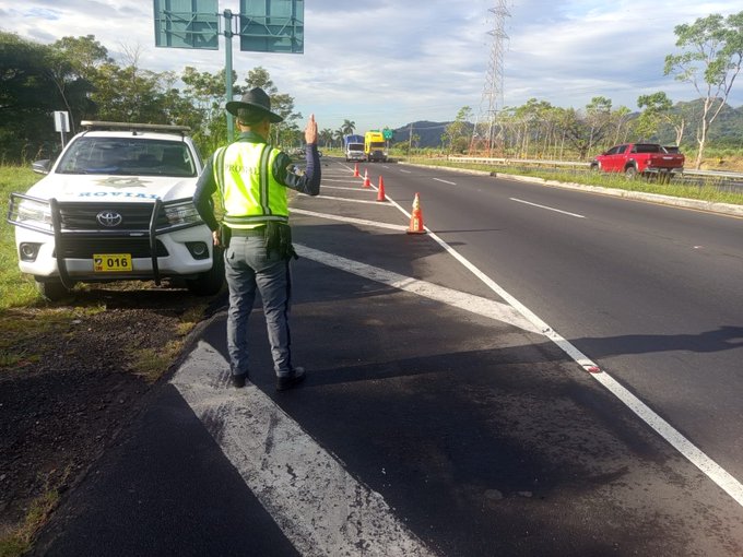 Un oficial de trafico con chaleco amarillo y sombrero esta de pie junto a un vehiculo Toyota blanco con luces de emergencia en una carretera asfaltada. Conos naranjas y barreras estan colocados en la via. Un camion y un vehiculo rojo son visibles en el fondo. Arboles y postes de energia se ven a los lados bajo un cielo nublado. Una senal de transito verde esta montada en un poste.