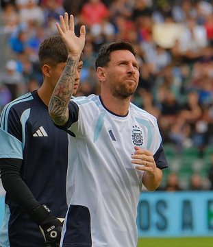 In a stadium filled with spectators, Lionel Messi stands on the field wearing a light blue and white Argentina national team jersey with the number 10 and Adidas logo, raising his right hand with fingers extended, his face showing a slight smile and focused expression, dark hair, next to a teammate in a dark blue sleeveless training top and shorts, with visible tattoos on the left arm, also raising a hand, both in athletic gloves, surrounded by blurred crowd in the background under stadium lights.
