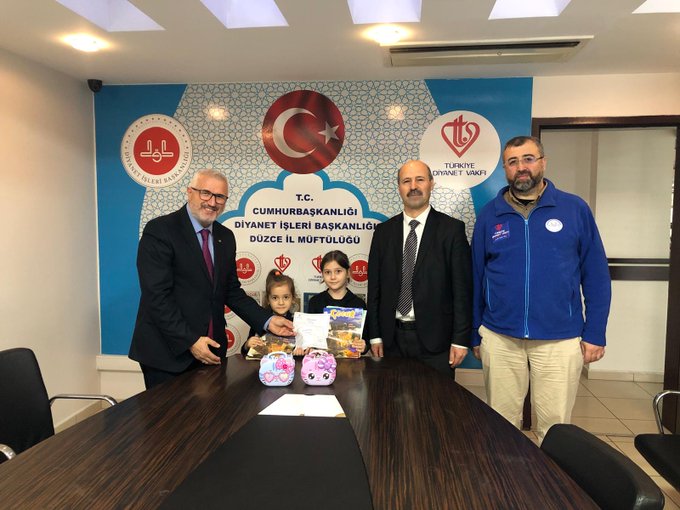 Two similar images show a group of four men in suits and jackets, two young girls in dark clothing, seated around a wooden conference table in a room with blue and white walls. The background features a large banner with Turkish flags, crescent moon symbols, and text reading CUMHURBAŞKANLIĞI DİYANET İŞLERİ BAŞKANLIĞI DÜZCE İL MÜFTÜLÜĞÜ. On the table are white papers, two pink gift bags with toys or books, and a rectangular pink item. One man holds a girls hand while another interacts with the second girl. The setting appears to be an official meeting for a charitable event.