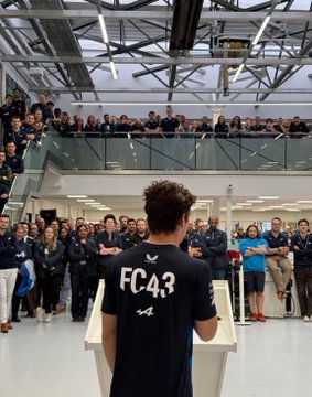 A young man with curly brown hair stands at a white podium in a modern industrial factory setting facing away from the camera wearing a black shirt with white FC43 lettering and Alpine F1 Team logo on the back surrounded by a diverse group of about 20-30 adults in dark suits and lanyards standing on the floor and upper levels clapping and watching attentively with bright overhead lighting and glass railings visible.