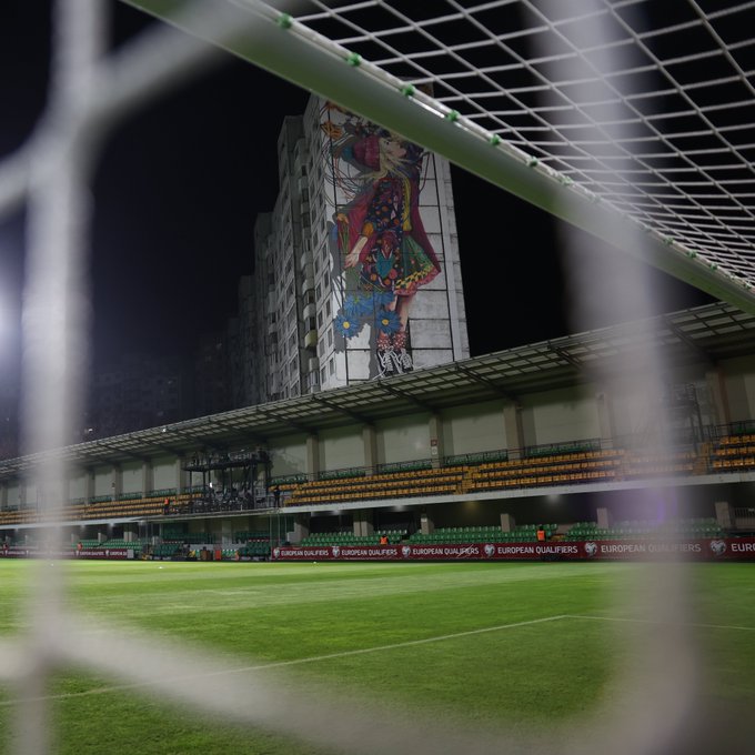 Nighttime view through football goalposts framing a green field and empty stadium stands with green seats. Large colorful mural on adjacent building depicts abstract figures in vibrant patterns. UEFA EURO 2024 logos on perimeter ads and banners around the pitch.