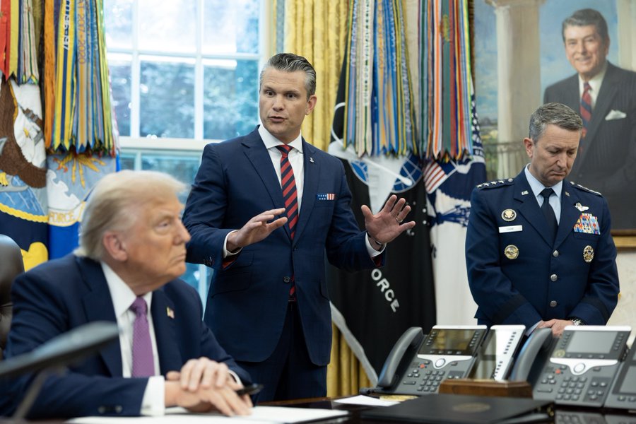 Em uma sala de reuniões formal, com bandeiras americanas e retratos, incluindo o de Ronald Reagan, nas paredes, o presidente Donald Trump, de terno escuro, senta-se à mesa gesticulando enquanto ouve atentamente; o secretário Pete Hegseth, de terno azul, está próximo, apontando; e um general em uniforme da Força Aérea observa atentamente ao lado de telefones e documentos sobre a mesa.