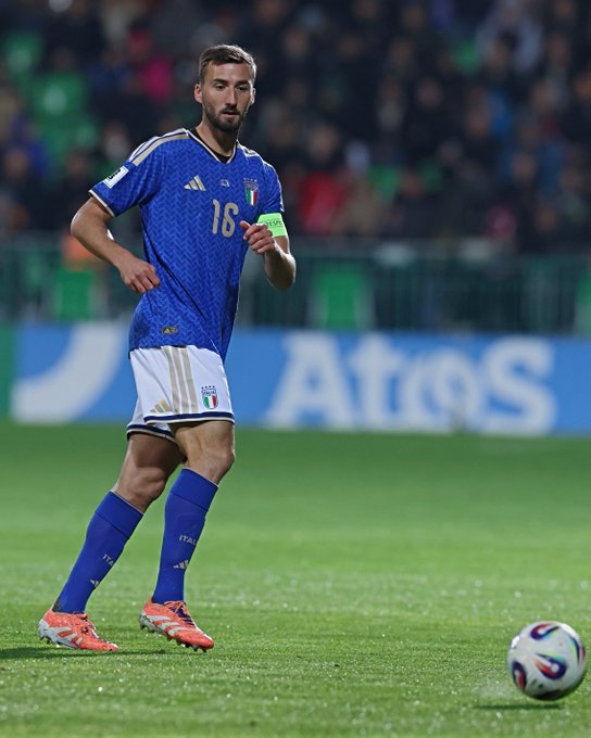 Bryan Cristante stands on a green soccer field in a blue Italy national team jersey with number 16 and green armband, white shorts featuring Italian flag patch, blue socks, and orange cleats. He positions to kick a colorful soccer ball nearby. Stadium seating with blurred crowd in green and blue attire fills the background. Atos sponsor board appears behind him.