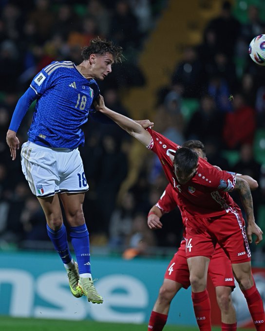 Italian soccer player in blue jersey number 18 jumps high with arms extended to head a white soccer ball mid-air during a match while a defender in red jersey number 24 grabs his arm attempting a tackle nearby players in similar uniforms crowd blurred in stadium stands yellow-lit field advertising boards visible at bottom.