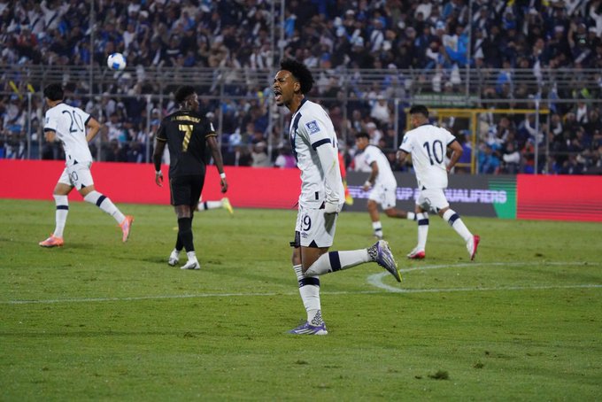 Soccer players in white and blue uniforms compete on a green field during a match with a scoreline visible on electronic boards showing Guatemala 1-2 Panama at the 69th minute players numbered 10 19 and 24 are active near the goal area with a referee in black and a crowded stadium in the background red barriers separate the field.
