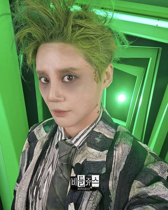 A person with bright green spiked hair and heavy black eye makeup around the eyes stands in a narrow green-lit hallway with multiple door frames. The individual wears a black and white striped suit jacket over a white shirt and striped tie. The face has pale skin and neutral expression. Another similar image shows the same person from a slightly different angle with the same costume and setting.