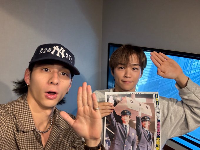 Two young men in casual attire one wearing a New York Yankees cap and checkered shirt with long hair and earrings posing with hands raised in salute next to a TV the other in light gray clothing with short hair also saluting while holding a printed photo of a uniformed stationmaster figure saluting against a plain wall background.