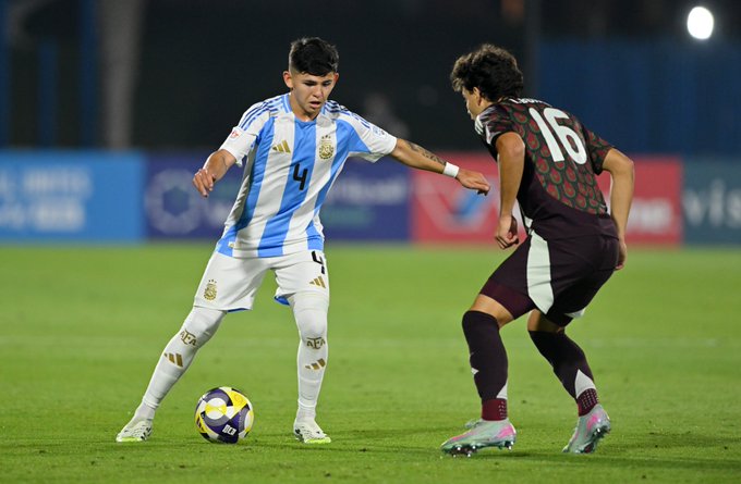 Two soccer players compete for the ball on a green field during a match under stadium lights. The player on the left wears Argentinas light blue and white striped jersey with number 4, white shorts, and white socks, kicking a yellow and green soccer ball. The player on the right wears a maroon and white jersey with number 16, maroon shorts, and maroon and pink socks, attempting to challenge. Blue advertising boards line the field background.