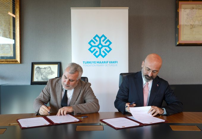 First image shows two men in suits, one with gray hair and glasses wearing a brown suit and tie, the other bald with a beard in a dark suit and pink tie, sitting at a wooden desk signing documents on red folders with Turkish crescent and star emblems, a white banner with blue Türkiye Maarif Vakfı logo in the background, framed photos on walls and a side table. Second image depicts the same two men standing side by side holding the red folders with emblems, smiling at the camera, against a similar banner setup in a formal room. Third image features six men in dark suits standing in a row in front of a wall with the Türkiye Maarif Vakfı logo and a Turkish flag, all appearing professional and posed for a group photo.