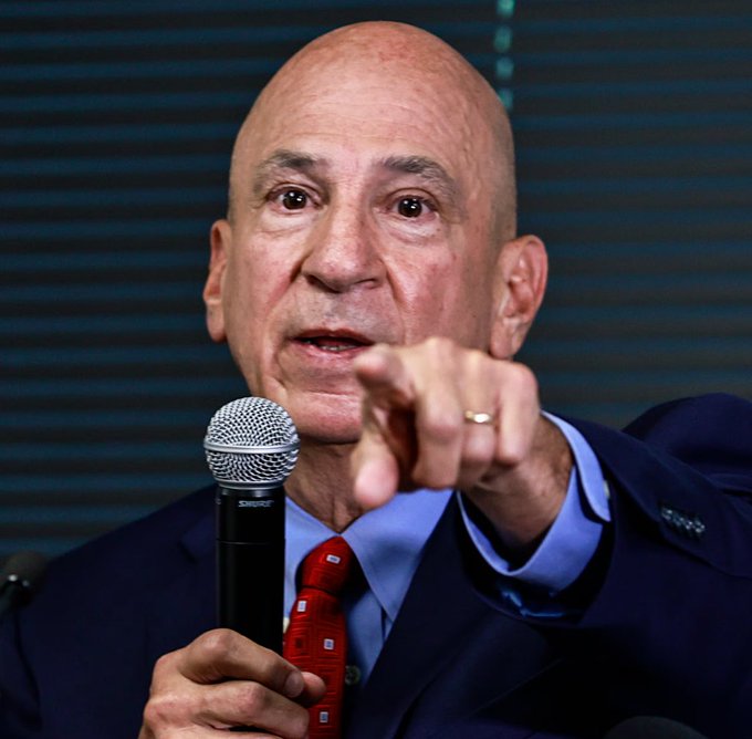A bald man with glasses wearing a blue suit and red tie stands at a podium holding a black microphone in his right hand while pointing forward with his index finger. He has a serious expression and is speaking. A wedding ring is visible on his left hand. The background features blurred vertical blinds and a wooden surface.