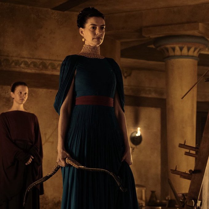 Anne Hathaway portrays Penelope wearing a teal pleated gown with flared sleeves gold necklace and red belt standing in an ancient hall holding a curved bow with another woman in maroon robe visible behind her. Tom Holland depicts Telemachus in bronze chest armor arm guards and skirt-like bottom holding a patterned clay bowl with a man in white robe blurred in background. Setting includes stone columns warm lighting hanging lamps wooden structures and pottery.