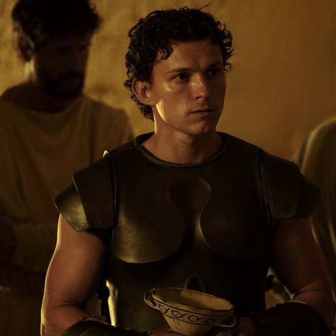 Anne Hathaway portrays Penelope wearing a teal pleated gown with flared sleeves gold necklace and red belt standing in an ancient hall holding a curved bow with another woman in maroon robe visible behind her. Tom Holland depicts Telemachus in bronze chest armor arm guards and skirt-like bottom holding a patterned clay bowl with a man in white robe blurred in background. Setting includes stone columns warm lighting hanging lamps wooden structures and pottery.