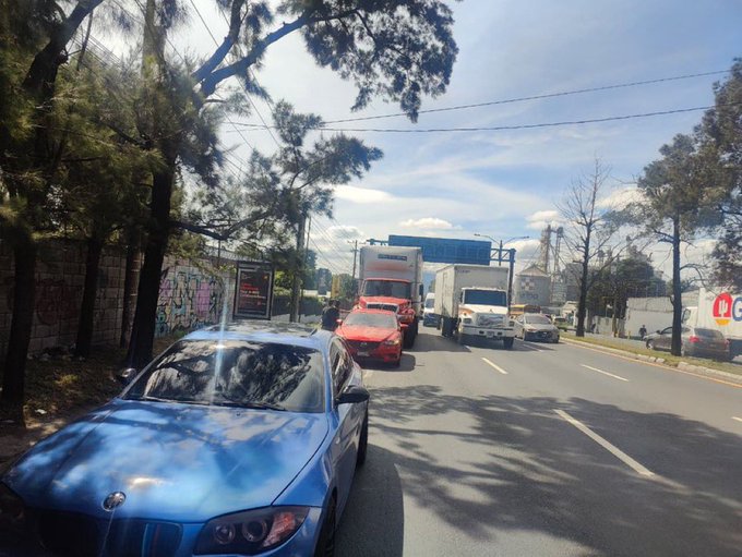A blue car positioned in the foreground on a multi-lane road with trees and a fence along the side under a partly cloudy sky. Behind it several vehicles including a red car multiple white trucks and a large semi-truck are stopped in traffic. A signboard is visible near the roadside amid the congestion suggesting an accident scene.
