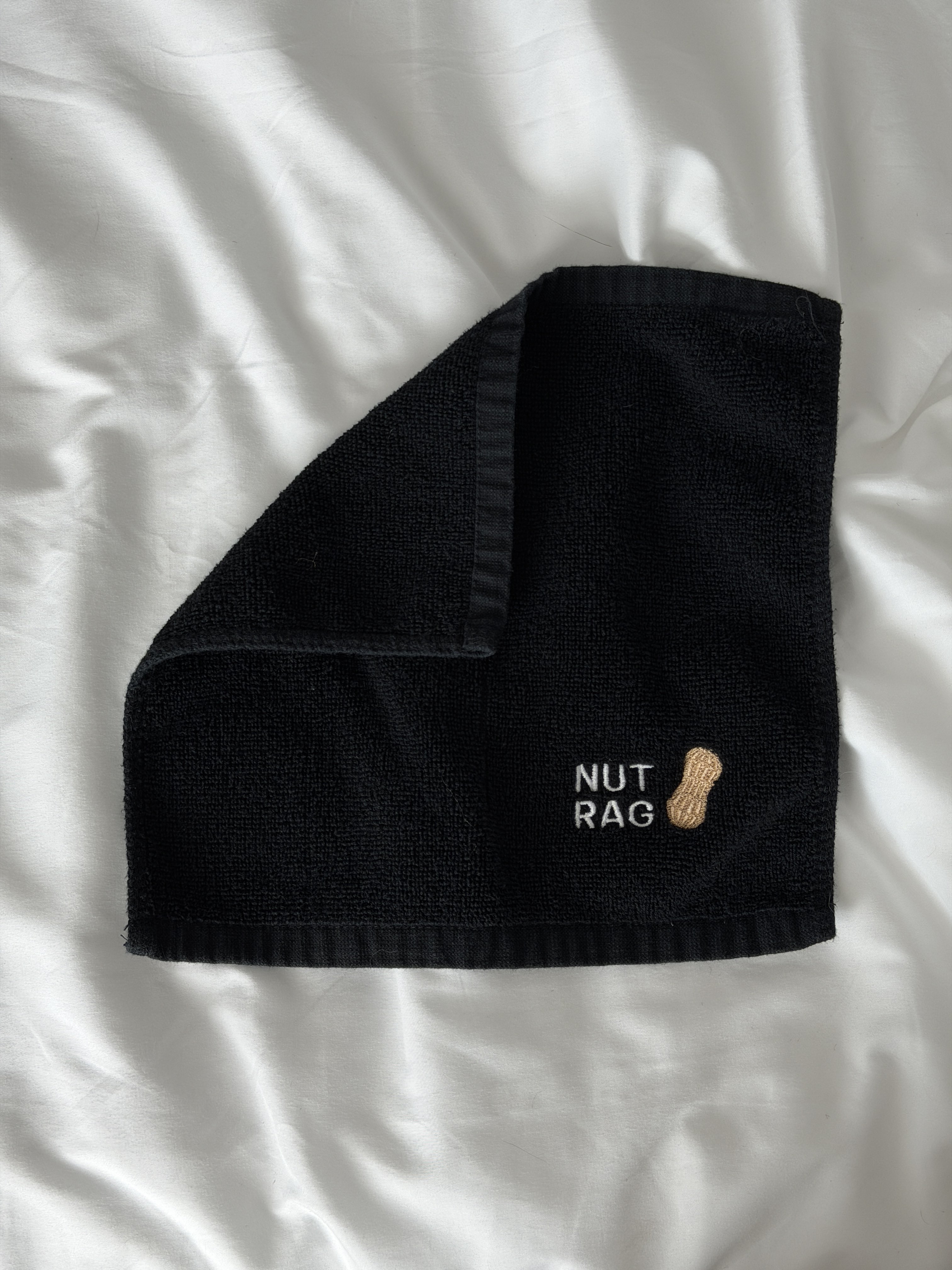 a black cumrag laying on a white bedsheet with an embroidered design in the bottom right corner that says NUT RAG with a peanut graphic.
