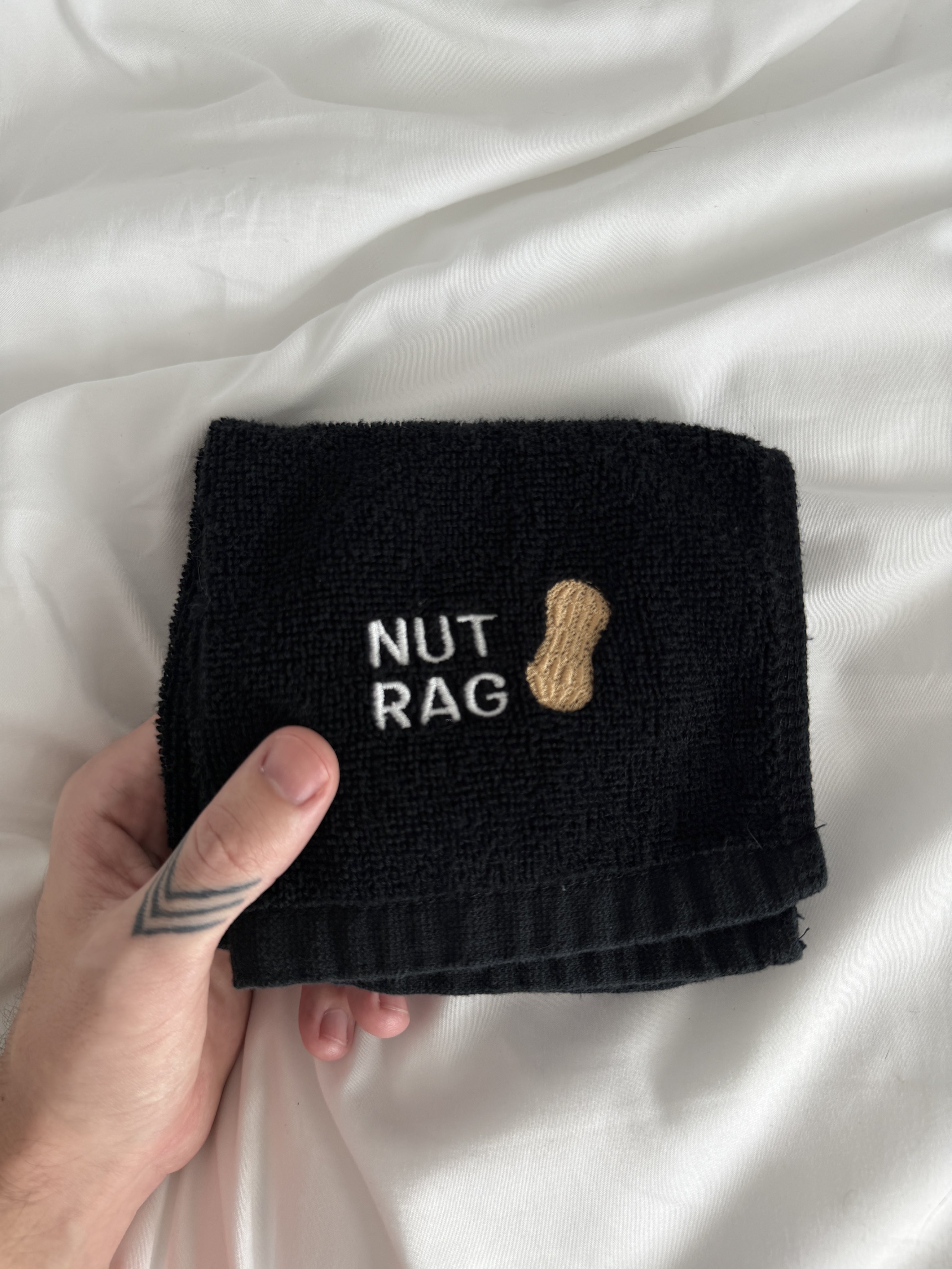a black cum rag with an embroidered design that says NUT RAG with a peanut graphic beside it, being held over a white bedsheet by a hand with a chevron thumb tattoo.