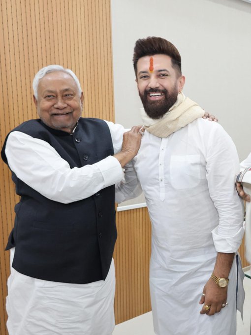 Two men standing side by side in a room with wooden paneling. The older man on the left wears a white kurta and black vest, smiling with hand on the younger mans shoulder. The younger man on the right wears a white kurta with beige scarf, smiling with hand on the older mans arm. Both have wristwatches and rings.