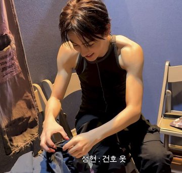 First image: Young man with short brown hair in black tank top and pants sits on white chair in room with blue walls, holding phone, examining small red circular marks on upper right arm, smiling slightly, bag on floor nearby. Second image: Same man in gray tank top leans forward over white bed in bedroom with window and nightstand, smiling at camera with playful expression, arms extended.