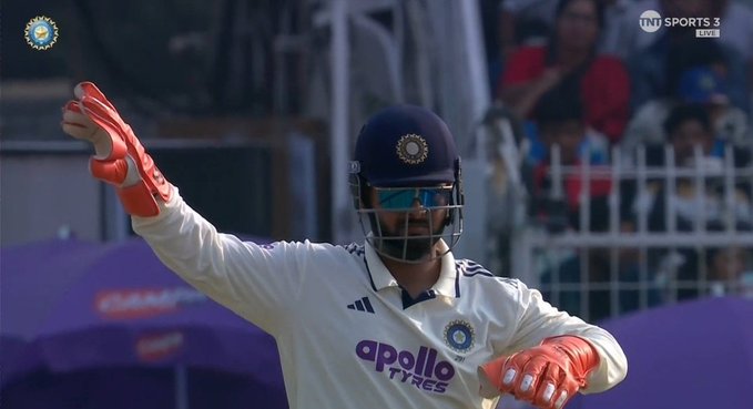 A male cricketer wearing a blue helmet with the Indian flag emblem, blue sunglasses, a white India jersey with Apollo sponsor logo, and red wicket-keeping gloves stands on a cricket field with his right arm extended forward and left arm slightly raised. He has a beard and is positioned near a purple umbrella and green netting in the background, with TNT Sports watermark visible.