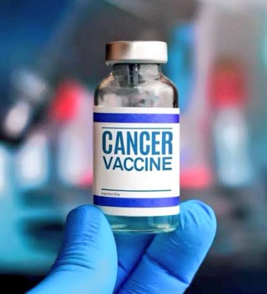 A clear glass vial containing a blue liquid, capped with a silver top, features a white label with blue stripes reading CANCER VACCINE in bold letters. The vial is held by a blue-gloved hand against a blurred blue and red background.