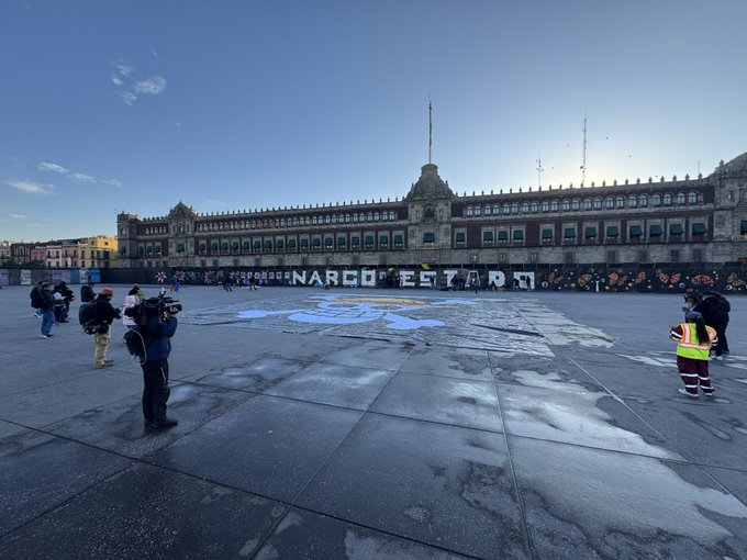 Large ornate historic building with multiple arches and towers serves as backdrop to expansive paved square under clear blue sky with setting sun. Prominent graffiti in white letters spells NARCOESTADO across the ground in front of the building. Several journalists with cameras and tripods positioned around the graffiti area. Few police officers in yellow vests stand nearby. Scattered people in casual and professional attire visible on the square.