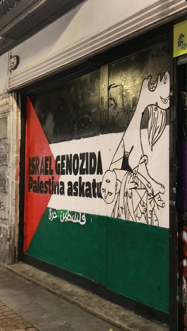 First image shows a poster on a wall with two soccer balls one in green and white stripes resembling Basque flag and another in black white green red resembling Palestinian flag text in Basque reads Elkartasuna Azaroa 17 Azarroak 15 Arrantzatu San Mames Era Herri libreak Euskal Herria Palestina Genozidio stop with logos at bottom including a green leaf and Bidetik text. Second image depicts graffiti on a building facade with Palestinian flag colors red black white green and text Israel Genozida in Basque a cartoonish figure with raised arms and Arabic script below.