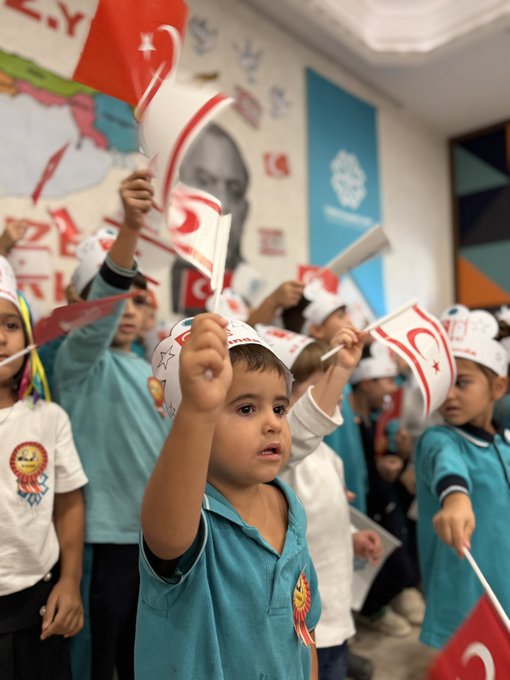 First image shows a group of school children in uniforms standing on a stage in a hall with a large mural of Mustafa Kemal Atatürk and the map of Northern Cyprus decorated with 42nd anniversary banners and flags waving Turkish and Northern Cyprus flags enthusiastically. Second image depicts young children wearing paper hats and holding white flags with red crescents and stars similar to Turkish flags raising them high in a classroom setting with wall decorations of flags and Atatürk portrait. Third image features children in blue uniforms wearing hats labeled KKTC and holding flags and making gestures in front of a wall with 42nd anniversary text and Northern Cyprus map elements. Fourth image captures four boys in turquoise uniforms sitting in an auditorium holding Turkish and Northern Cyprus flags smiling at the camera with some wearing badges.