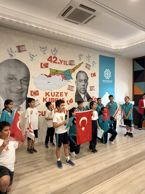 First image shows a group of school children in uniforms standing on a stage in a hall with a large mural of Mustafa Kemal Atatürk and the map of Northern Cyprus decorated with 42nd anniversary banners and flags waving Turkish and Northern Cyprus flags enthusiastically. Second image depicts young children wearing paper hats and holding white flags with red crescents and stars similar to Turkish flags raising them high in a classroom setting with wall decorations of flags and Atatürk portrait. Third image features children in blue uniforms wearing hats labeled KKTC and holding flags and making gestures in front of a wall with 42nd anniversary text and Northern Cyprus map elements. Fourth image captures four boys in turquoise uniforms sitting in an auditorium holding Turkish and Northern Cyprus flags smiling at the camera with some wearing badges.