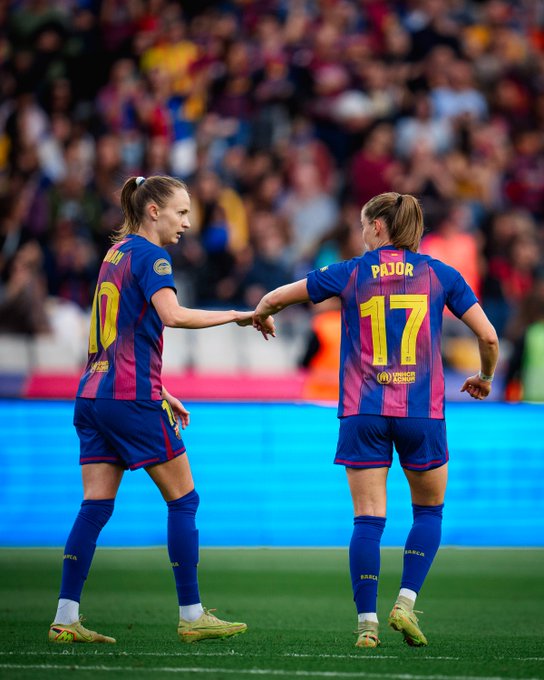 Two women in blue and red FC Barcelona Femení soccer jerseys and shorts, one with ponytail and number 10 on back, the other with ponytail and number 17 named Pajor on back, stand on green grass field shaking hands with right hands, wearing yellow cleats, in front of blue stadium barrier and blurred crowd spectators in stands.