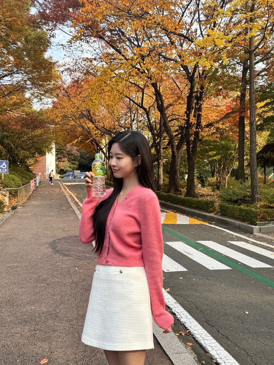 Kinju's tweet image. I wandered around a park glowing with autumn colors, letting myself unwind a little. In my soft pink cardigan and white skirt, I sat by the benches with a drink in hand, enjoying the breeze and the quiet moment I didn’t know I needed.